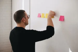 A man adding a sticky note to a row of notes on a white wall, appearing focused and engaged in planning or organization.