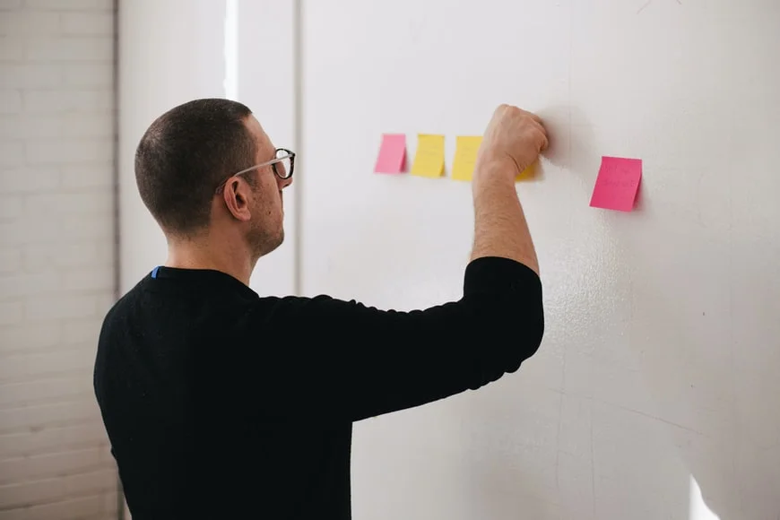 A man adding a sticky note to a row of notes on a white wall, appearing focused and engaged in planning or organization.